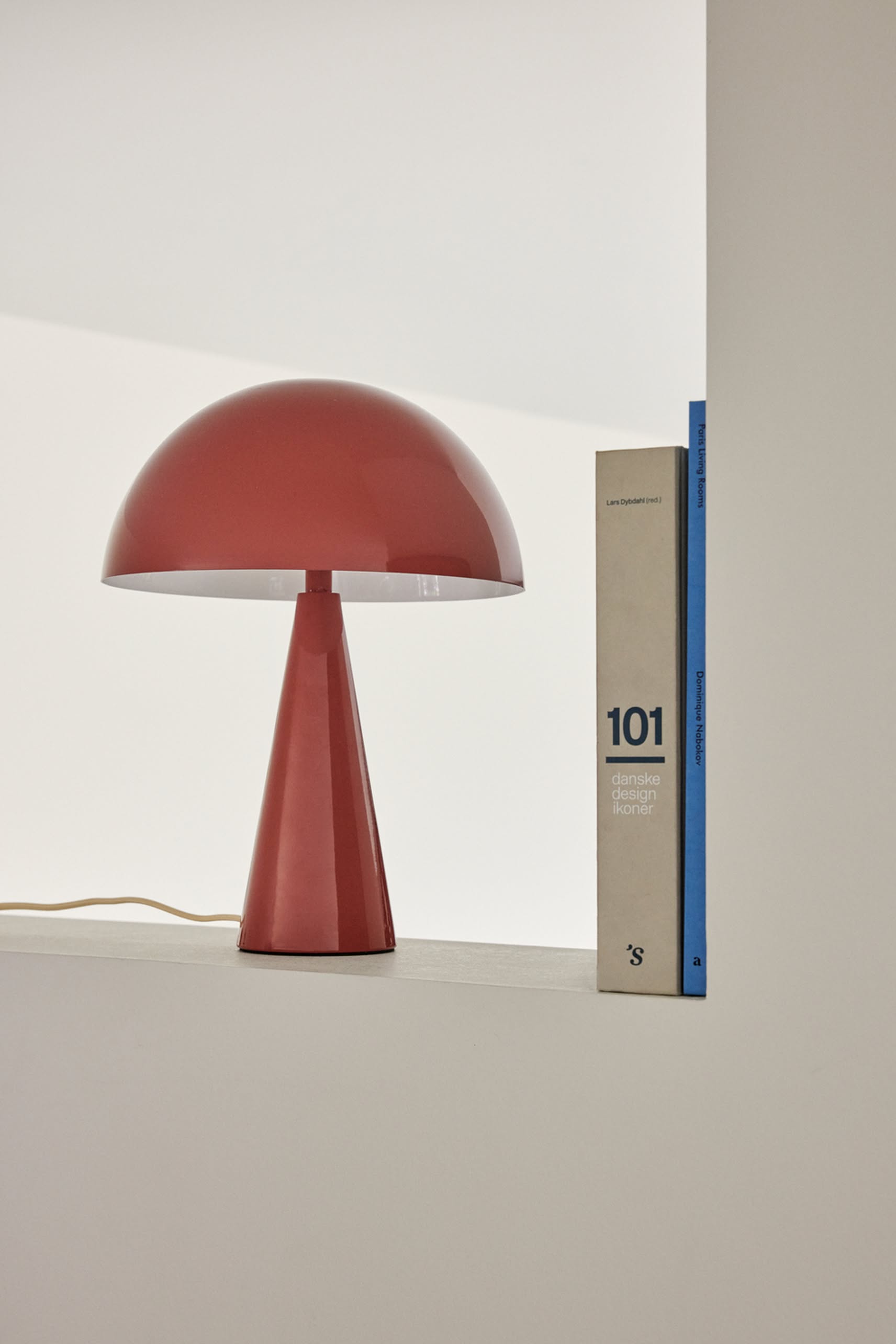 A Hübsch Mush Table Lamp Mini with a red dome shade from Hübsch is displayed on a white shelf beside the book 101 danske designikoner—a stylish accent for modern homes, offered in multiple colors.