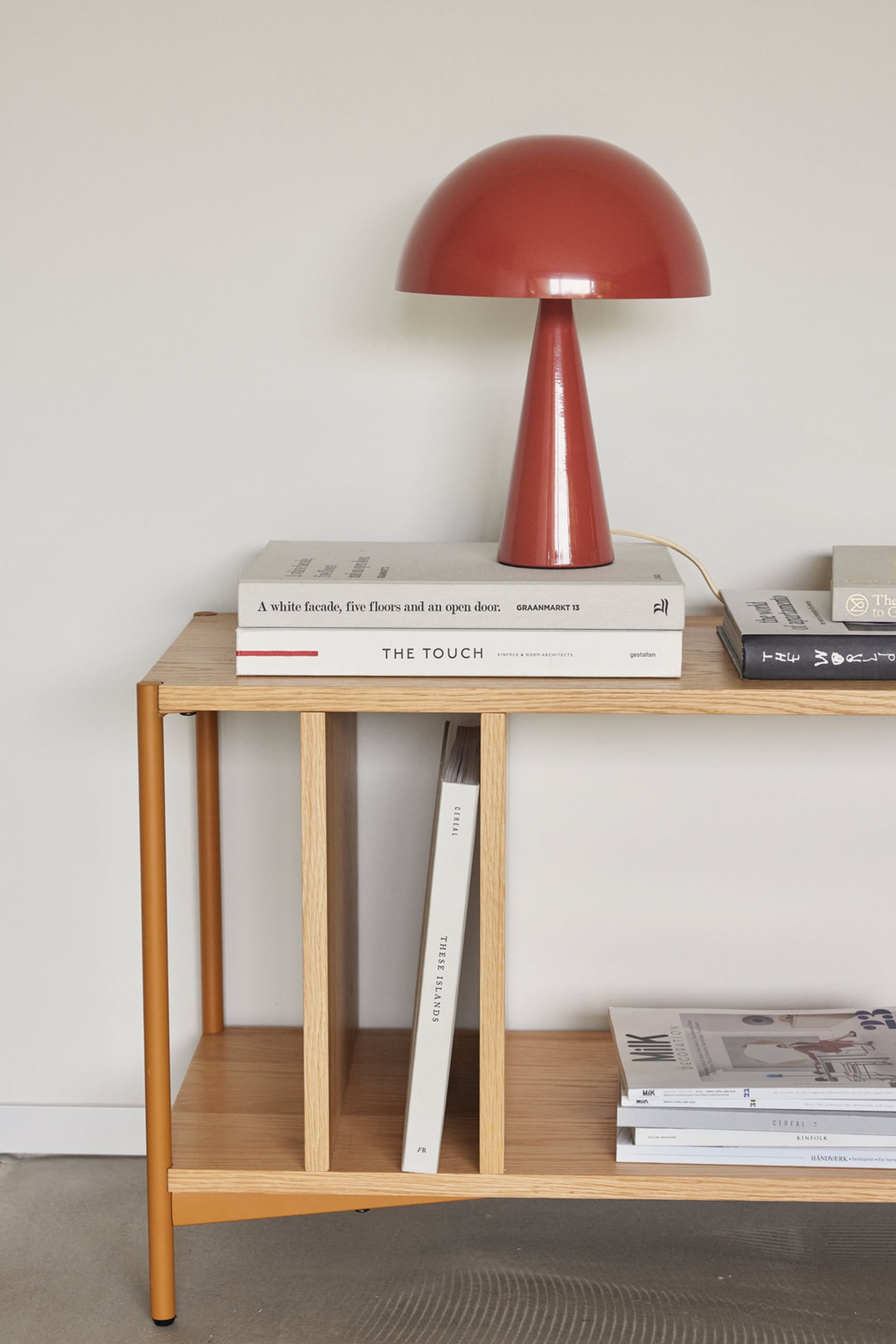 A modern wooden side table with orange legs displays stacked books and a red Hübsch Mush Table Lamp Mini by Hübsch. More books and magazines are on the lower shelf, set against a plain white wall—perfect for any modern home.
