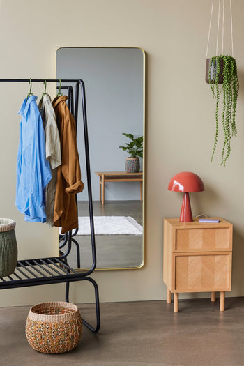 A modern entryway features a black coat rack with shirts, a basket, a large wall mirror, two potted plants, and the Hübsch Mush Table Lamp Mini in red by Hübsch on a wooden side table beside a notepad.
