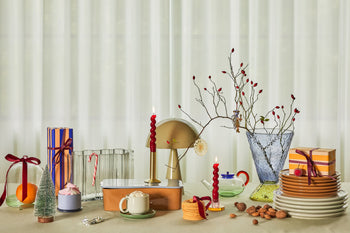 A festive table features wrapped gifts, colorful glassware, desserts, nuts, twisted red candles, the Hübsch Mush Table Lamp Mini in assorted colors by Hübsch, and a clear vase with branches—ideal for modern homes with sheer white curtains.