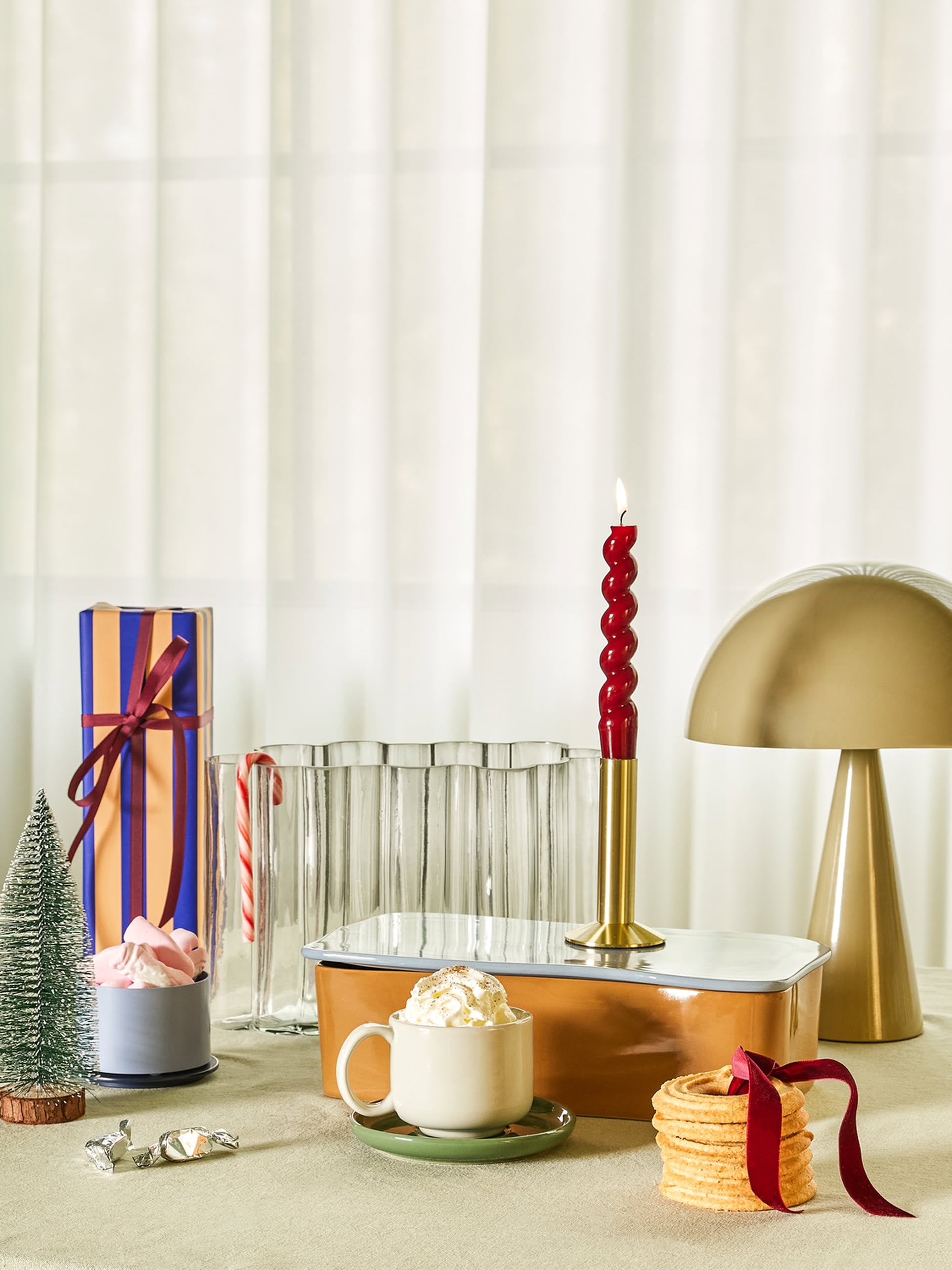 A festive table showcases the Hübsch Mush Table Lamp Mini by Hübsch, a mug of whipped cream-topped hot chocolate, waffle cookies, wrapped gifts in various colors, a red candle, a candy cane, and mini Christmas trees—ideal for the modern home.