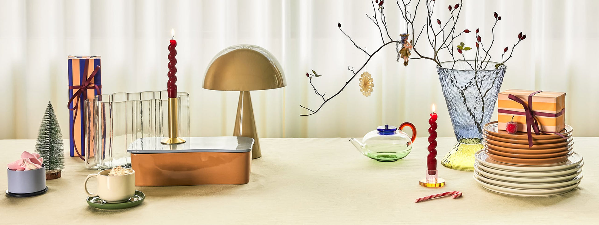 A festive table set with plates, wrapped gifts, cocoa, candles, a Hübsch Mush Table Lamp Mini in various colors, a teapot, and a glass vase with branches creates the perfect modern home vibe against sheer white curtains.