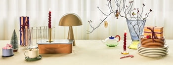 A festive table set with plates, wrapped gifts, cocoa, candles, a Hübsch Mush Table Lamp Mini in various colors, a teapot, and a glass vase with branches creates the perfect modern home vibe against sheer white curtains.