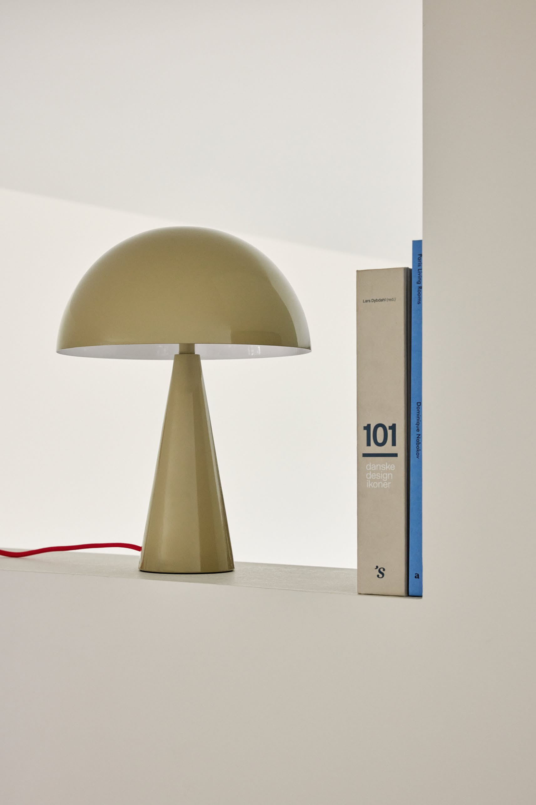 A Hübsch Mush Table Lamp Mini in beige, accented by a red cord, sits beside a large white book titled 101 Danish Design Icons on a minimalist white surface—an ideal touch of style for any modern home.