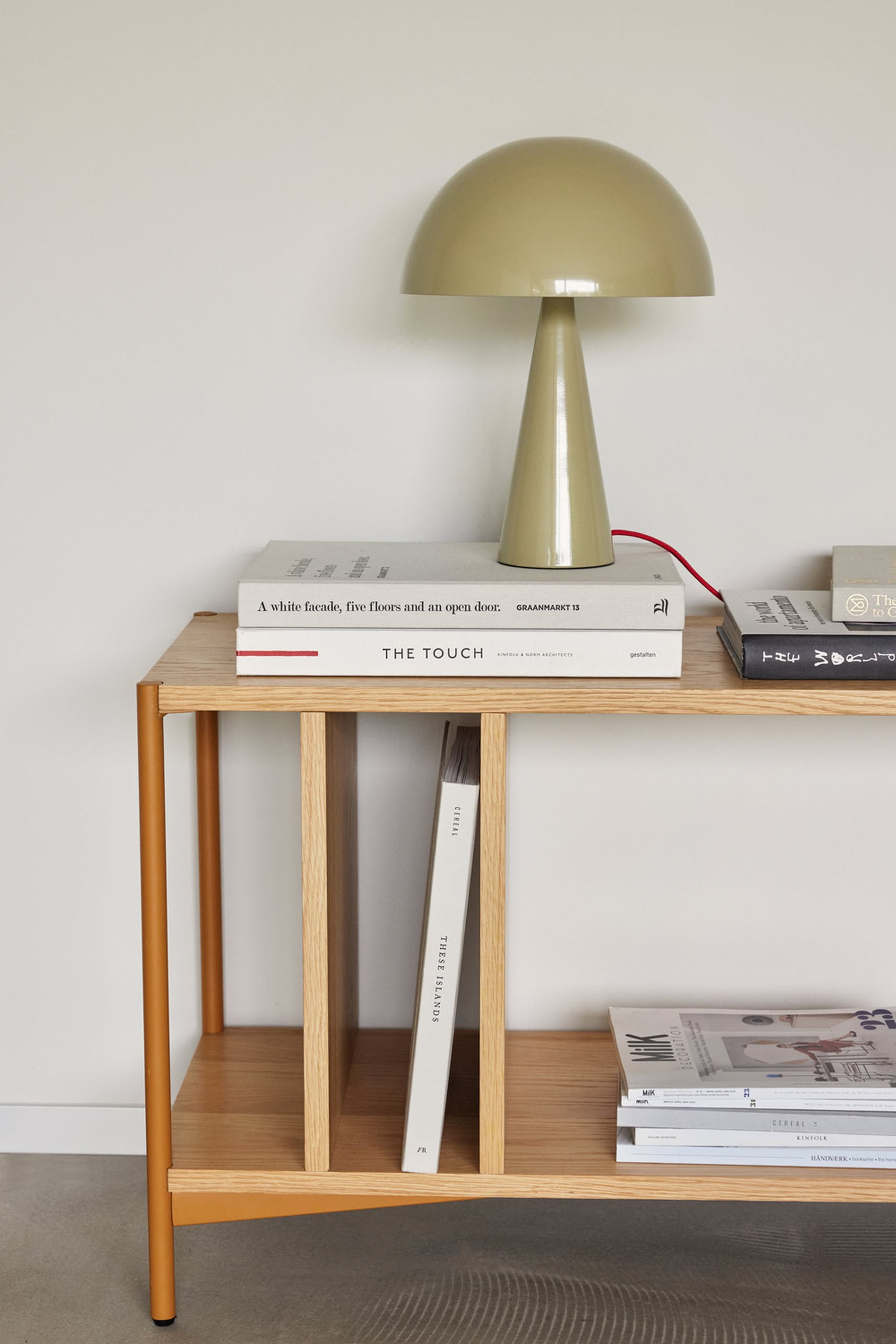 A modern wooden side table displays the beige Hübsch Mush Table Lamp Mini and stacked books, with more books and magazines on lower shelves against a plain white wall—ideal for a stylish home, with various color options available.