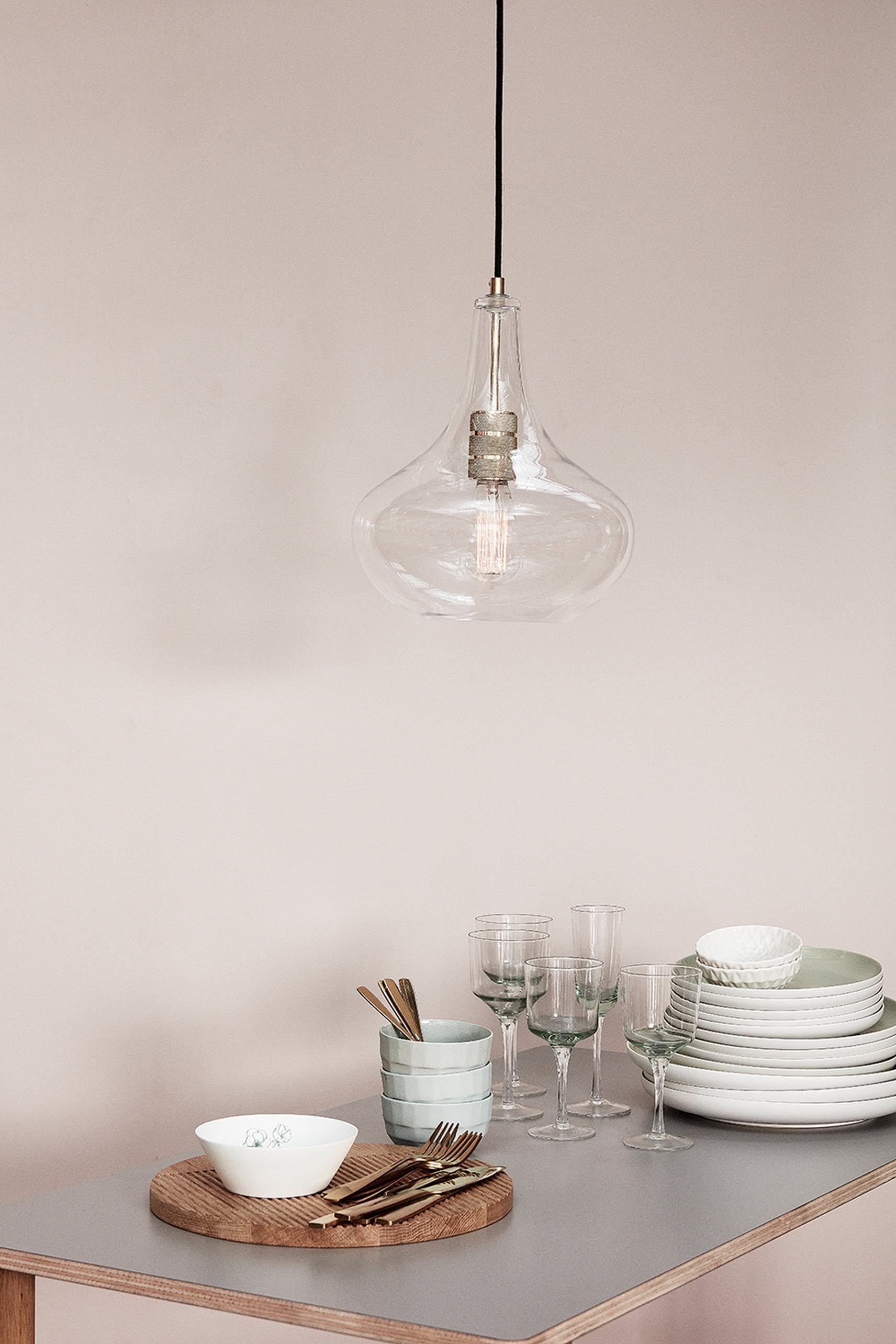A clear glass pendant light hangs above a table set with versatile kitchenware, featuring the Hübsch Select Bowls, Set of 5 by Hübsch, alongside stacked white dishes, glassware, and gold cutlery on a wooden board against a soft beige wall.