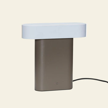 The Hübsch Sleek Table Lamp by Hübsch features a rectangular brown base, a flat oval white top, and a black power cord, creating a modern minimalistic look against a plain background.