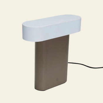 The Hübsch Sleek Table Lamp by Hübsch features an oval white top, a rectangular metallic brown base, and a black power cord, offering a modern minimalistic look that stands out against light backgrounds.