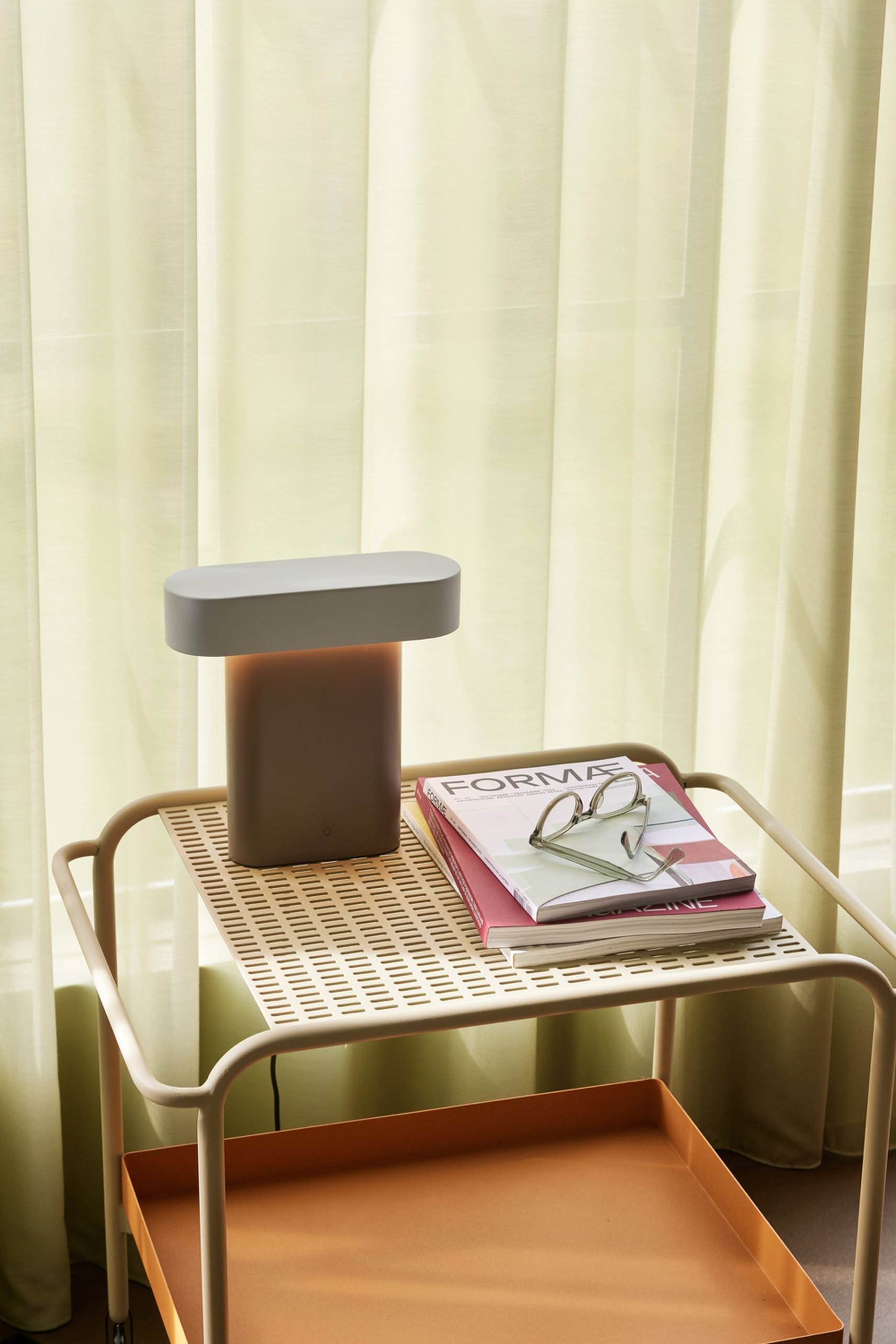 A small beige side table holds the Hübsch Sleek Table Lamp by Hübsch with dimmable function, a pair of glasses, and two books including FORMA. Sheer curtains in the background softly diffuse natural light for a modern minimal look.
