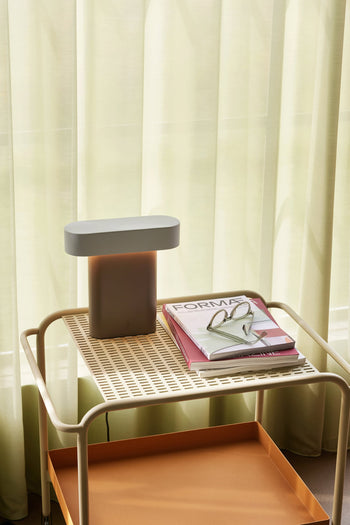 A small beige side table holds the Hübsch Sleek Table Lamp by Hübsch with dimmable function, a pair of glasses, and two books including FORMA. Sheer curtains in the background softly diffuse natural light for a modern minimal look.