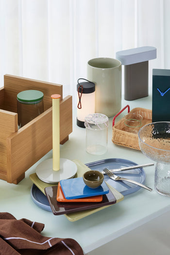 A table set with colorful plates, bowls, cutlery, glass jars, a wicker basket, wooden box, ceramic vases, decorative objects, and the Hübsch Sleek Table Lamp by Hübsch—all arranged before a white curtain.