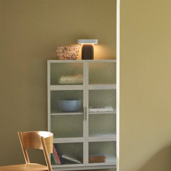 A wooden chair sits beside a glass-front cabinet. On top are stacked items and the Hübsch Sleek Table Lamp by Hübsch, featuring a dimmable function that enhances the modern minimalistic style against the beige wall.