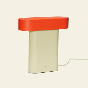 The Hübsch Sleek Table Lamp by Hübsch features a modern minimalistic design with a rectangular beige base, flat oval red top, and power cord, shown against an off-white background.