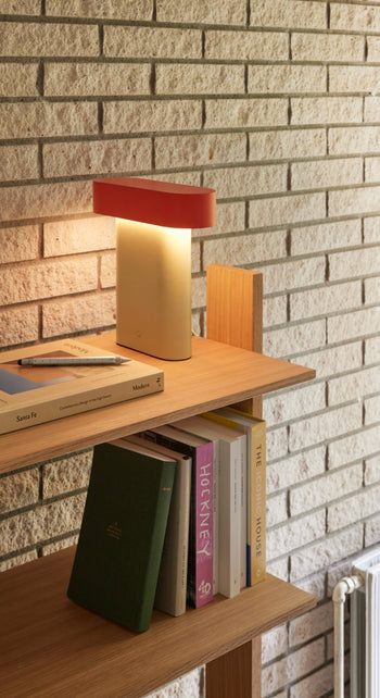 A wooden shelf against a light brick wall holds books, a closed notebook with a pen, and the Hübsch Sleek Table Lamp by Hübsch with a red shade and soft yellow glow, creating a modern minimalistic vibe in the space.