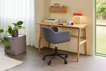 A modern home office features a wooden desk, gray swivel chair, and the Hübsch Sleek Table Lamp by Hübsch with dimmable function. Books and papers are on the desk, shelves above, and a plant sits by the window with white curtains.