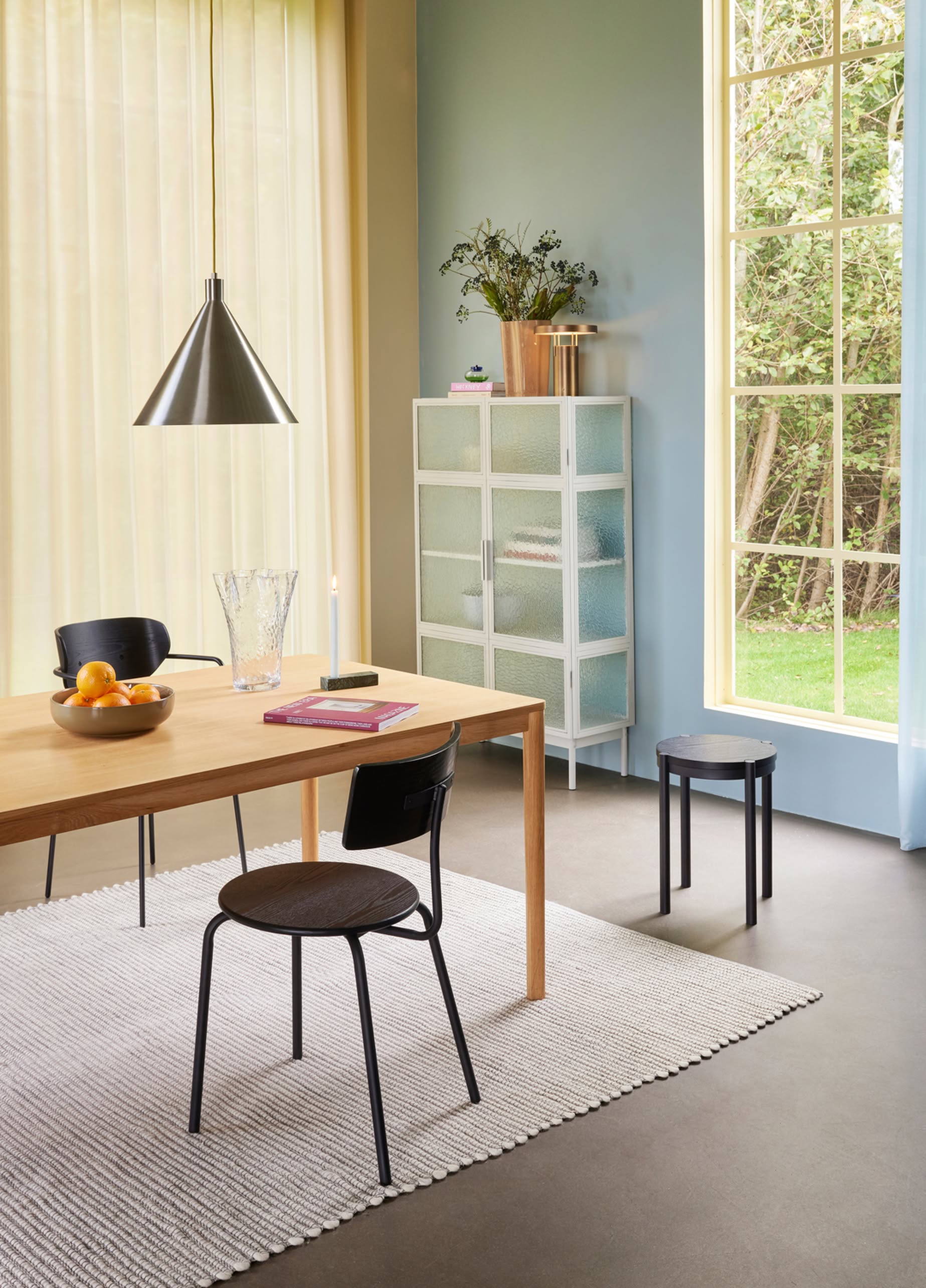 A modern dining area with a wooden table, black chairs, and fruit bowl exudes elegance. Sunlight highlights the striped rug, white cabinet with plants, and the Hübsch Yama Pendant by Hübsch hanging overhead.