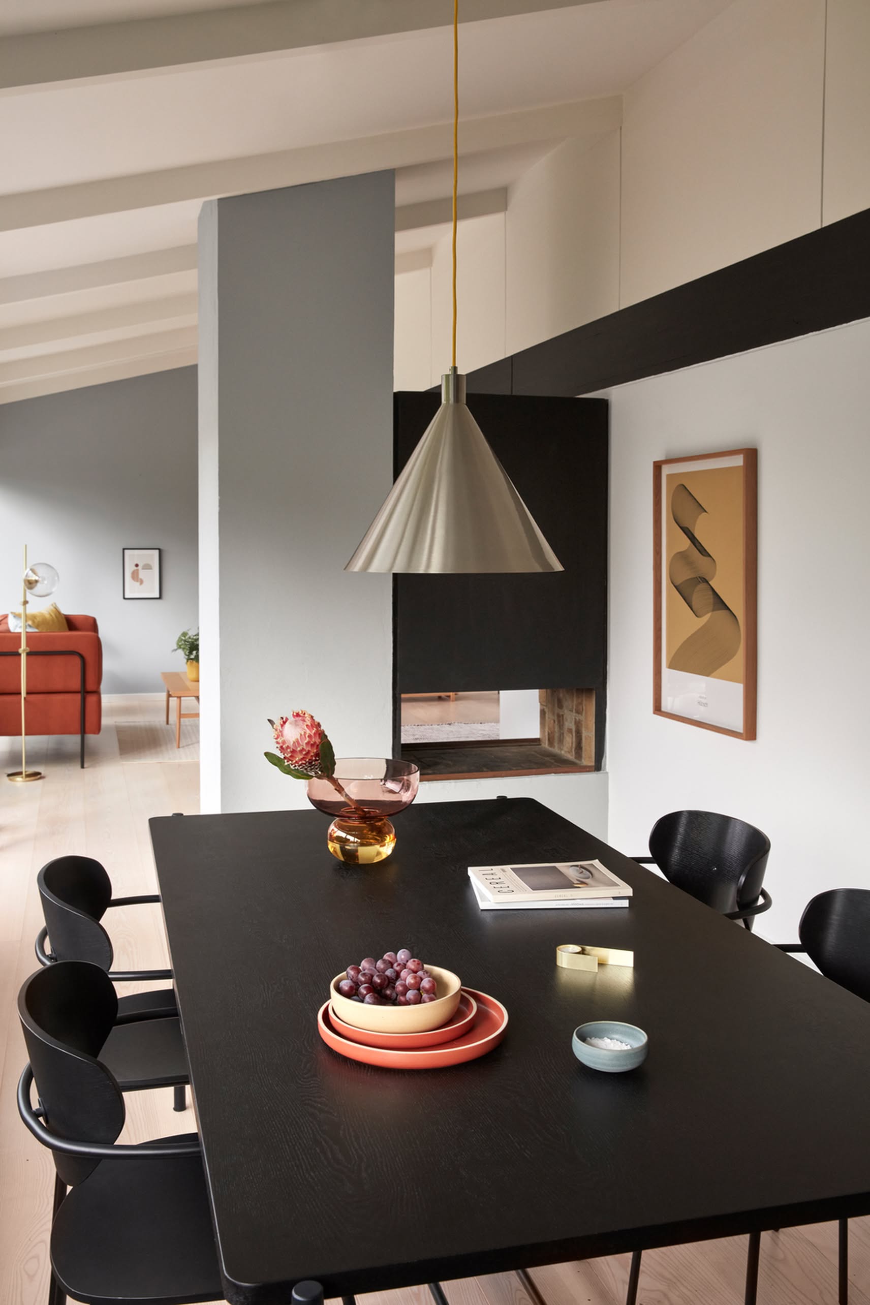 A modern dining room exudes elegance with a black table, sleek chairs, fruit bowls, books, and a flower vase. The striking Hübsch Yama Pendant hangs above, art decorates the wall, and a red sofa adds a pop of color in the background.