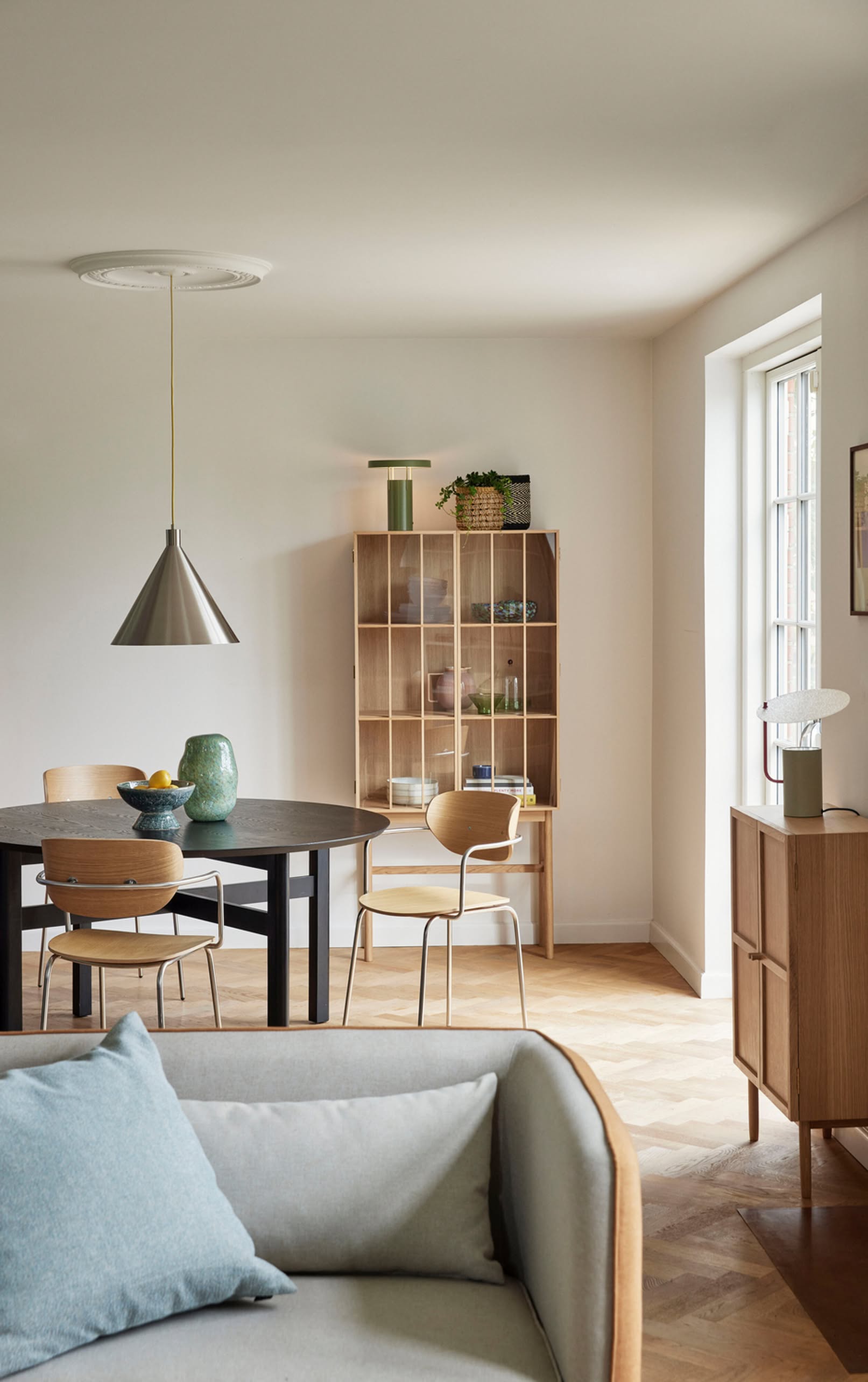 A modern dining room featuring a round black table, four wooden chairs, and a glass-front cabinet with plants and decor. Natural light highlights the wooden sideboard, while the Hübsch Yama Pendant by Hübsch brings modern elegance overhead.