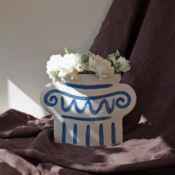 The INI CERAMIQUE Greek Column Ceramic Vase by INI CERAMIQUE, featuring blue patterns, displays white flowers atop a brown cloth as sunlight casts elegant shadows.