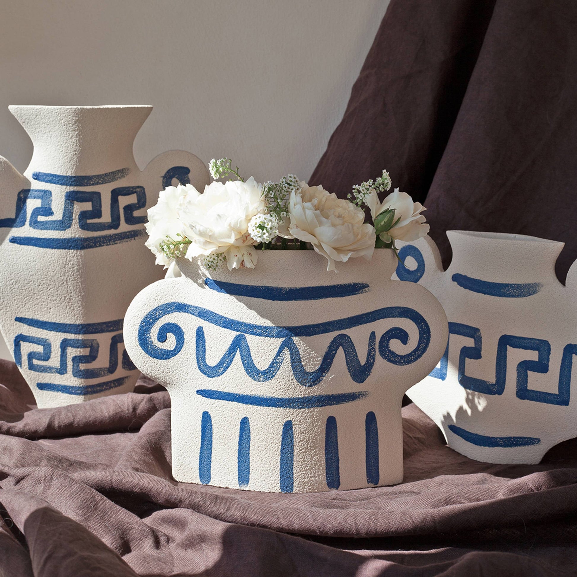 Three textured ceramic vases, including the INI CERAMIQUE Greek Column Ceramic Vase by INI CERAMIQUE with blue Greek patterns, sit on a brown cloth. A unique vase holds white flowers as soft light creates gentle shadows on the scene.