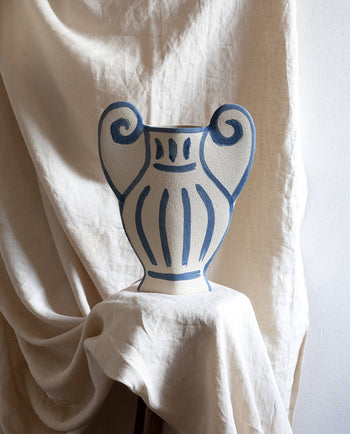 The INI CERAMIQUE Krater N°1 Ceramic Vase, featuring blue painted patterns and two handles, is displayed on a cream-draped stool against a softly lit, textured backdrop—blending Greek pottery tradition with modern design.