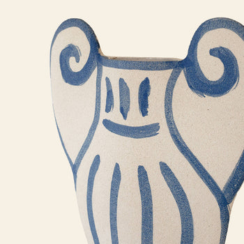The INI CERAMIQUE Krater N°1 Ceramic Vase features a cream base with blue painted designs—smiling face and swirls—blending Greek pottery influences with modern style, all set against a light beige background.