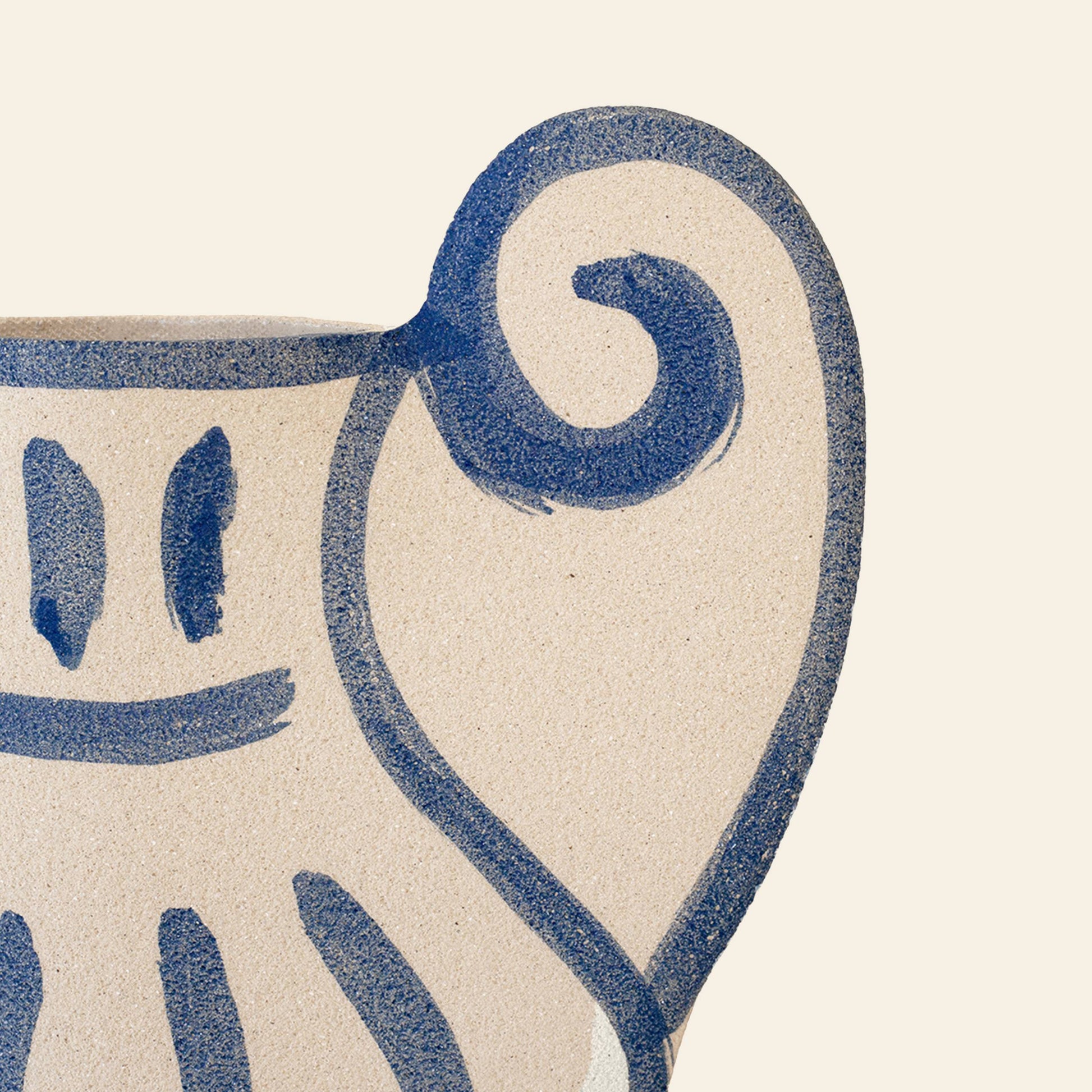 A close-up of the INI CERAMIQUE Krater N°1 Ceramic Vase, a beige piece featuring blue abstract lines and facial motifs, merges Greek-inspired pottery with modern design. The spiral handle detail contrasts beautifully against the plain light background.
