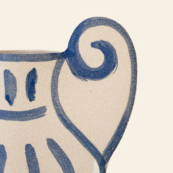 A close-up of the INI CERAMIQUE Krater N°1 Ceramic Vase, a beige piece featuring blue abstract lines and facial motifs, merges Greek-inspired pottery with modern design. The spiral handle detail contrasts beautifully against the plain light background.