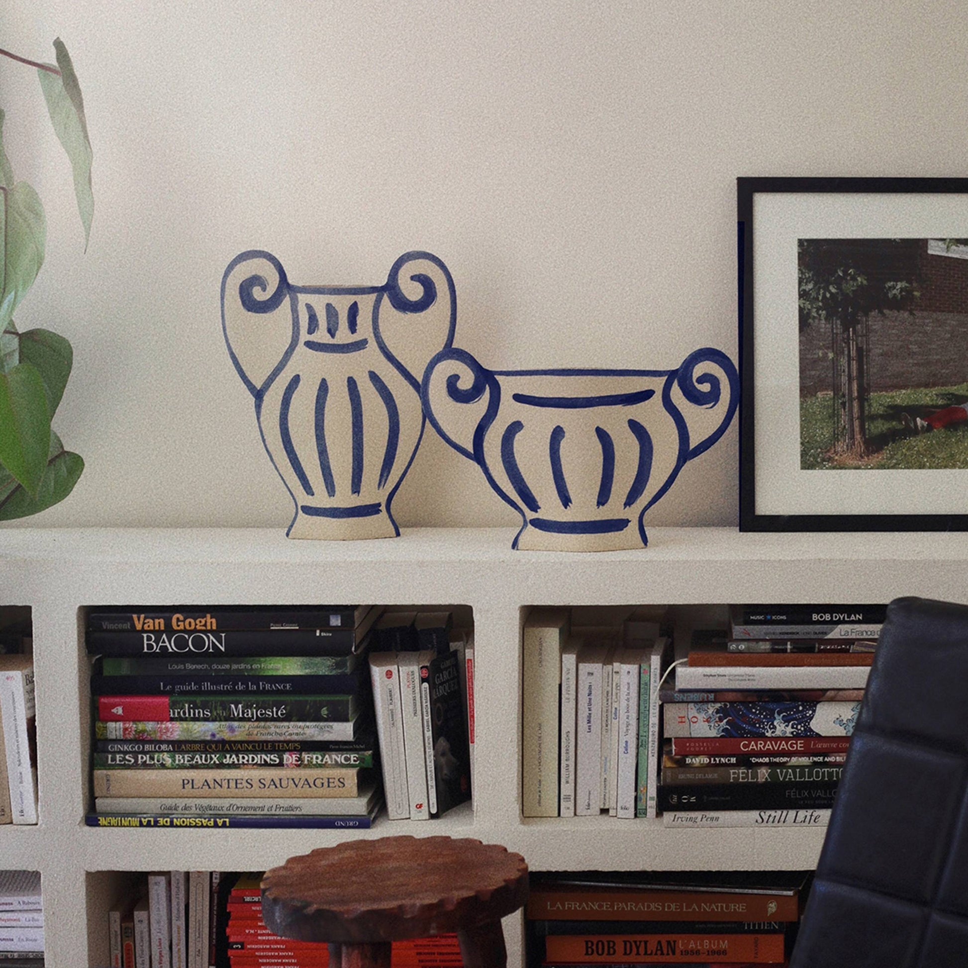 A white bookshelf with art books, an INI CERAMIQUE Krater N°1 Ceramic Vase by INI CERAMIQUE, a framed photo, a green plant on the left, and a black chair in front create a contemporary design vibe.