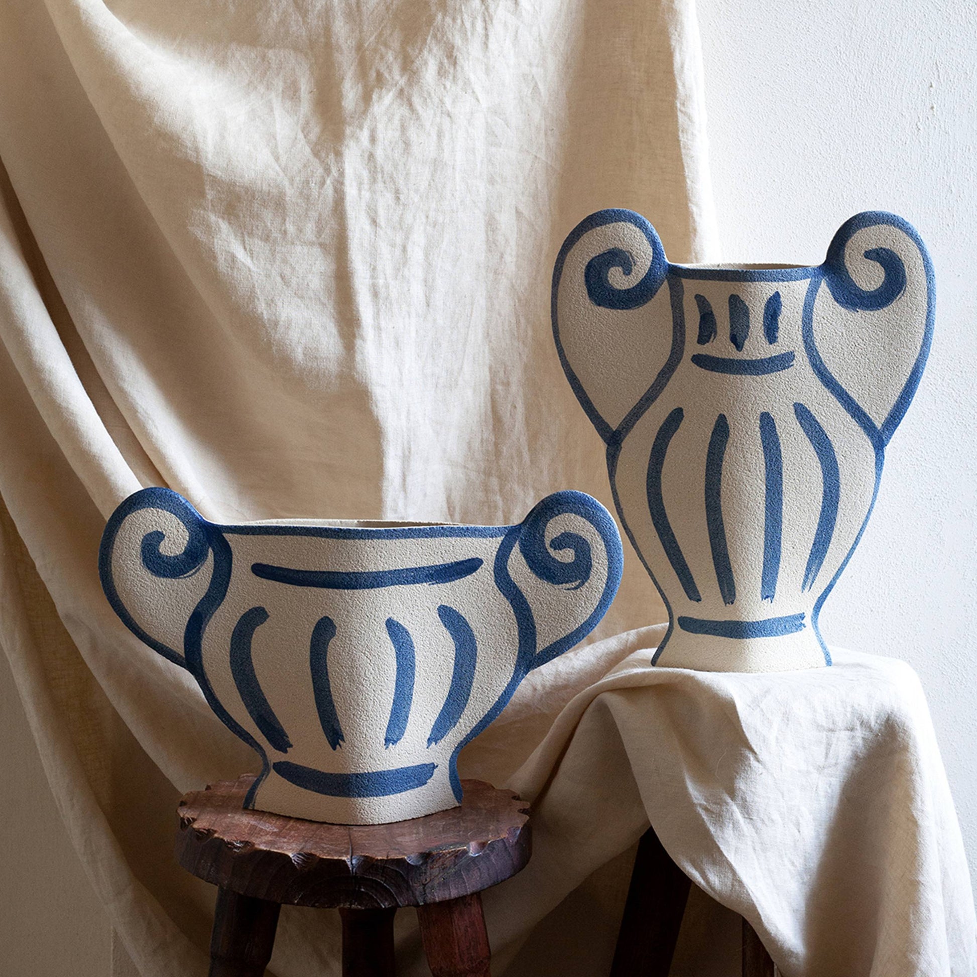 Two INI CERAMIQUE Krater N°1 Ceramic Vases with blue Greek-inspired patterns and handles are showcased on wooden stools draped in beige fabric, set against a beige textured backdrop.