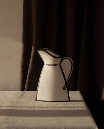 The INI CERAMIQUE Morandi Pitcher Ceramic Vase with black outlines rests on a beige cloth-covered table, its artisanal craftsmanship accentuated by dramatic light and shadows against a softly lit wall and dark curtain backdrop.