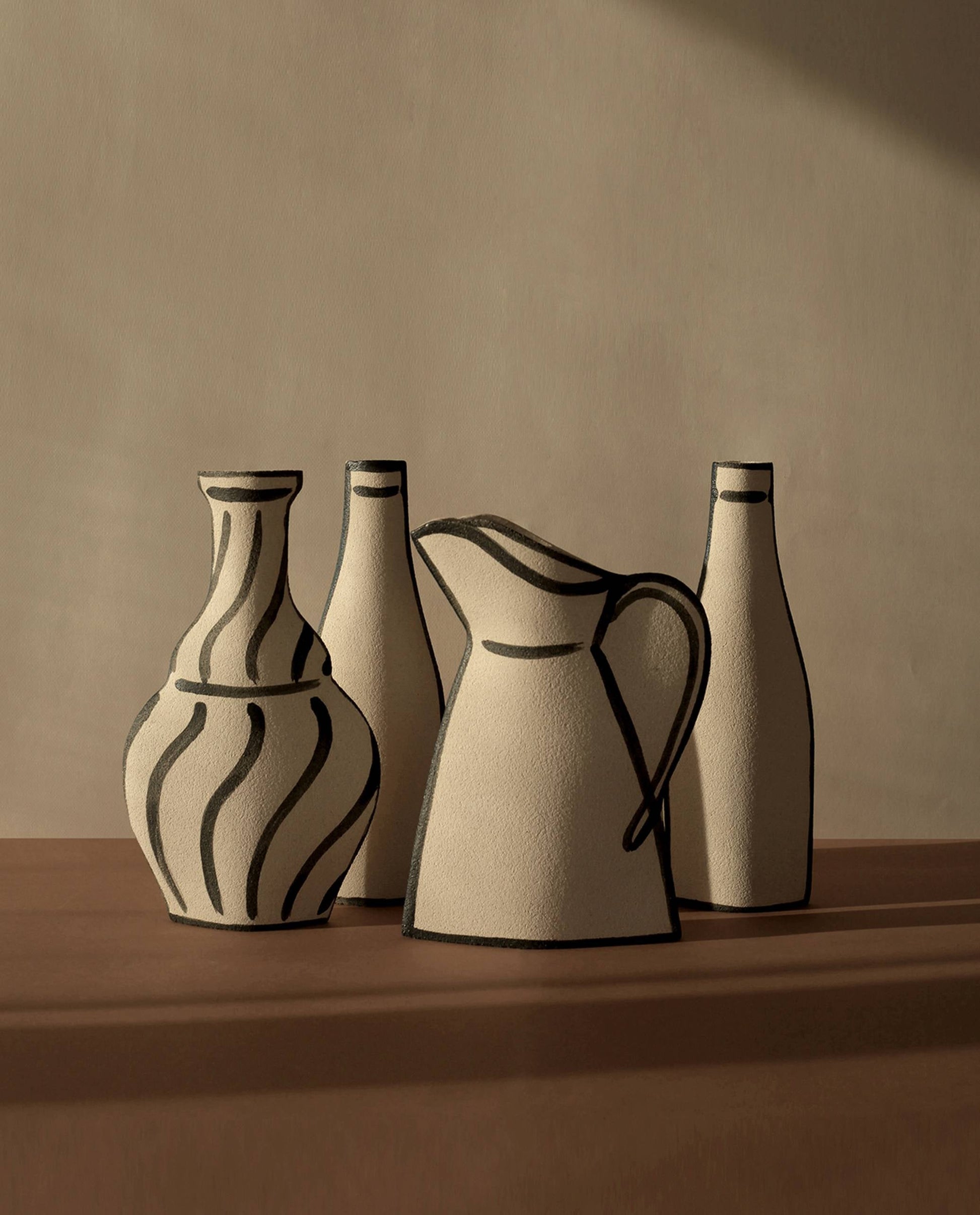 A minimal set of four beige ceramic vessels by INI CERAMIQUE—including the Morandi Pitcher Ceramic Vase and two bottles with bold black outlines—is arranged on a brown surface, highlighting artisanal craftsmanship.