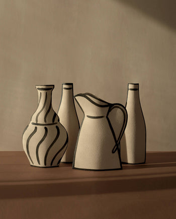 A minimal set of four beige ceramic vessels by INI CERAMIQUE—including the Morandi Pitcher Ceramic Vase and two bottles with bold black outlines—is arranged on a brown surface, highlighting artisanal craftsmanship.