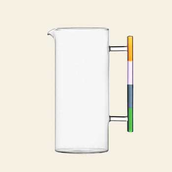 The Ichendorf Milano Andalusia Jug is a clear, handmade borosilicate glass pitcher with a cylindrical shape, straight spout, and a colorblocked gradient handle in stacked yellow, orange, purple, blue, and green.
