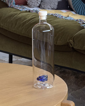 An Ichendorf Milano Animal Farm Bottle Big by Ichendorf Milano, featuring a small blue glass bear inside and a white cap, sits empty on a wooden table. A green couch draped with a colorful blanket is visible in the background.