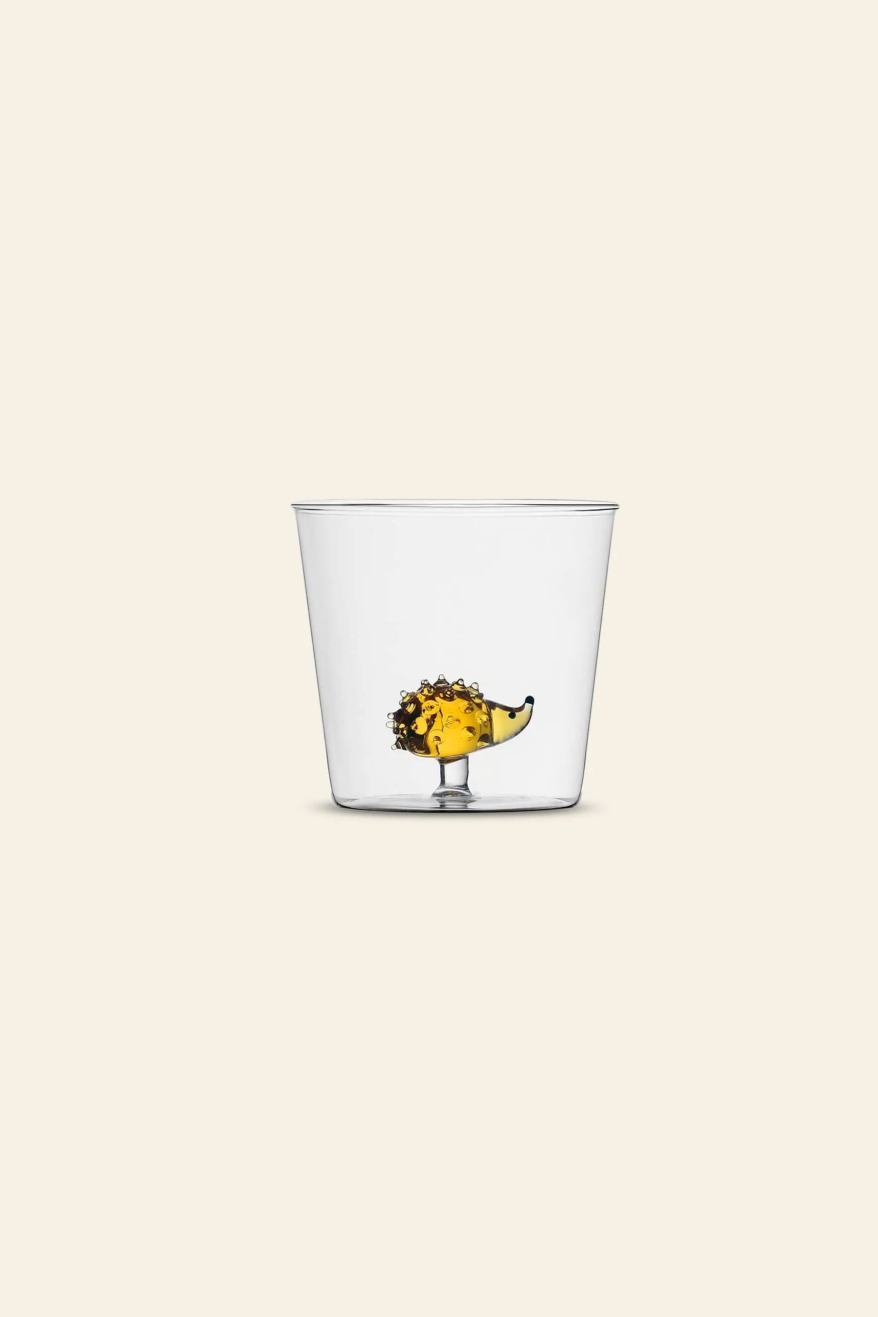 The Ichendorf Milano Animal Farm Tumbler by Ichendorf Milano is a clear glass featuring a handcrafted hedgehog-shaped base inside, holding yellow liquid, on a light beige background, making it look as if the liquid fills the hedgehog.