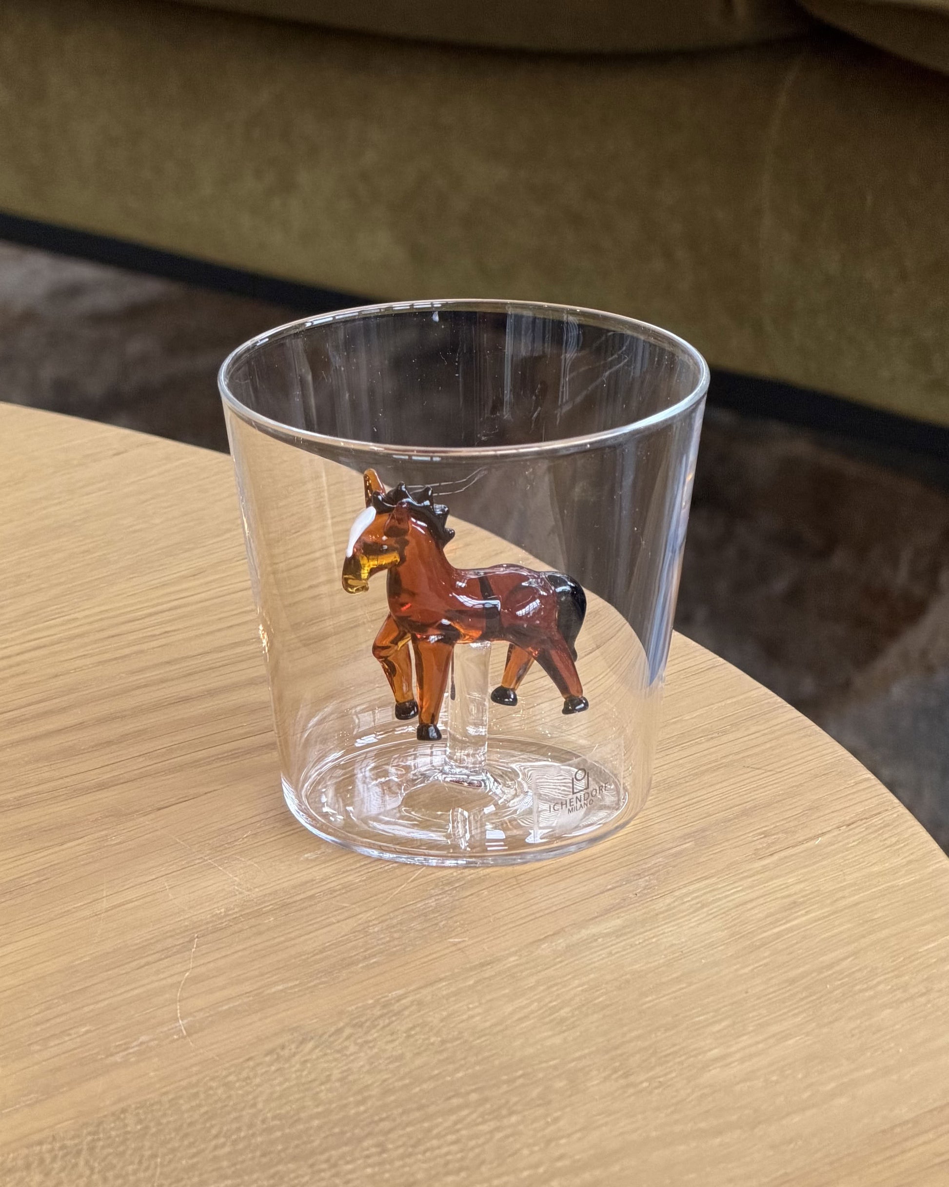 An Ichendorf Milano Animal Farm Tumbler rests on a wooden table, its base featuring a brown glass horse. In the background are a green couch and brown rug.