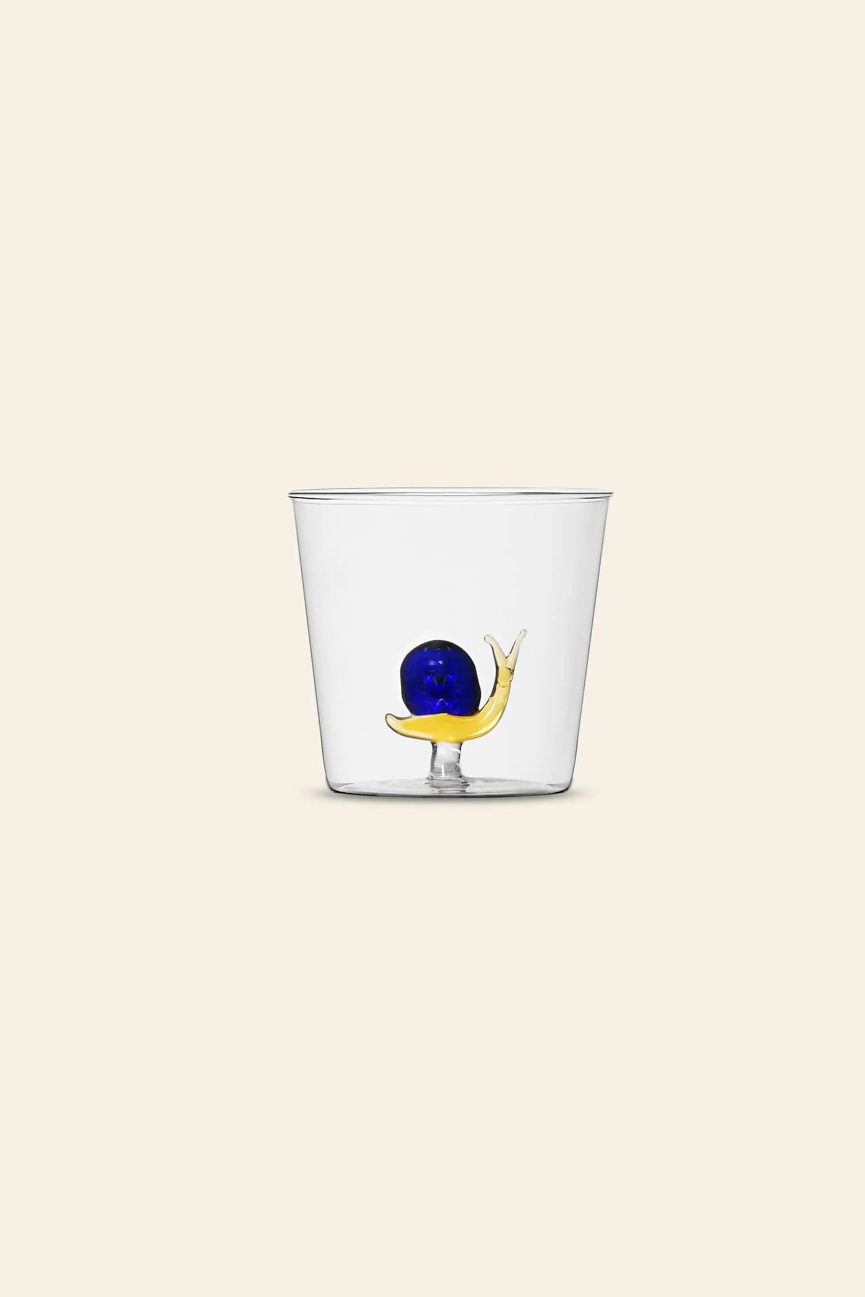 The Ichendorf Milano Animal Farm Tumbler by Ichendorf Milano is a clear glass cup featuring a whimsical yellow and blue snail inside, showcasing handcrafted charm, photographed against a plain beige background.