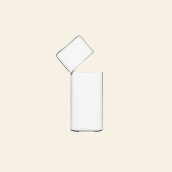 Two clear Ichendorf Milano Attesa Flower Vases (Medium) are balanced—one upright, the other tilted and resting on its rim with unique charm—against a plain, light beige background.