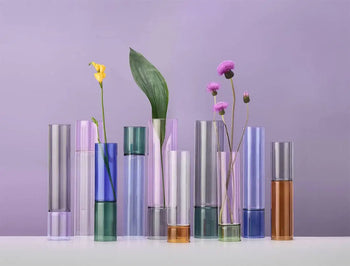 A row of Ichendorf Milano Bamboo Groove Vases (27cm) holds levitating single blooms and leafy stems, displayed on a white surface with a light purple background.