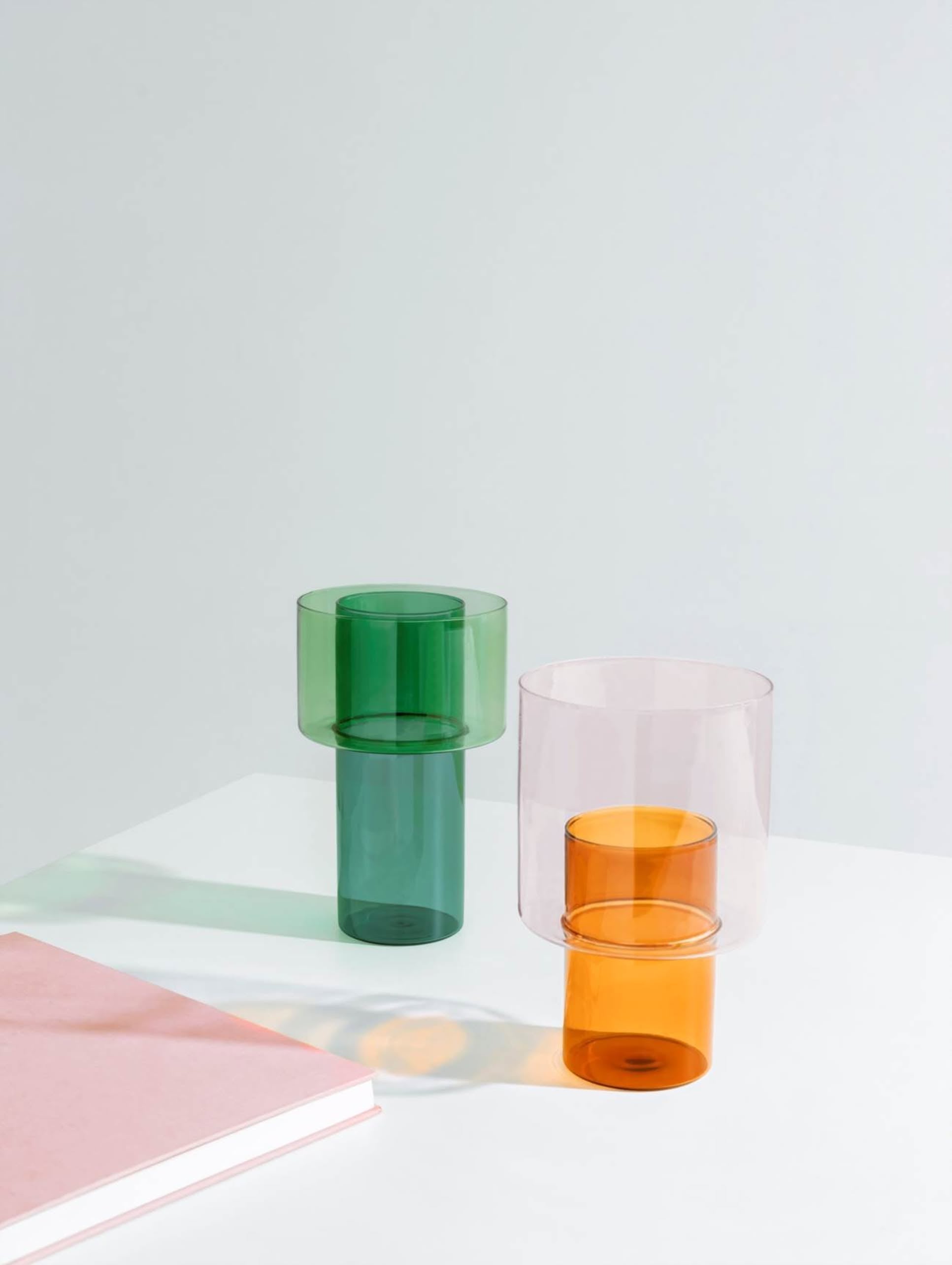 The Ichendorf Milano Boreale Vase A by Ichendorf Milano, in translucent green and orange, rests on a white surface near a pink book, casting colorful shadows against a plain light gray backdrop.