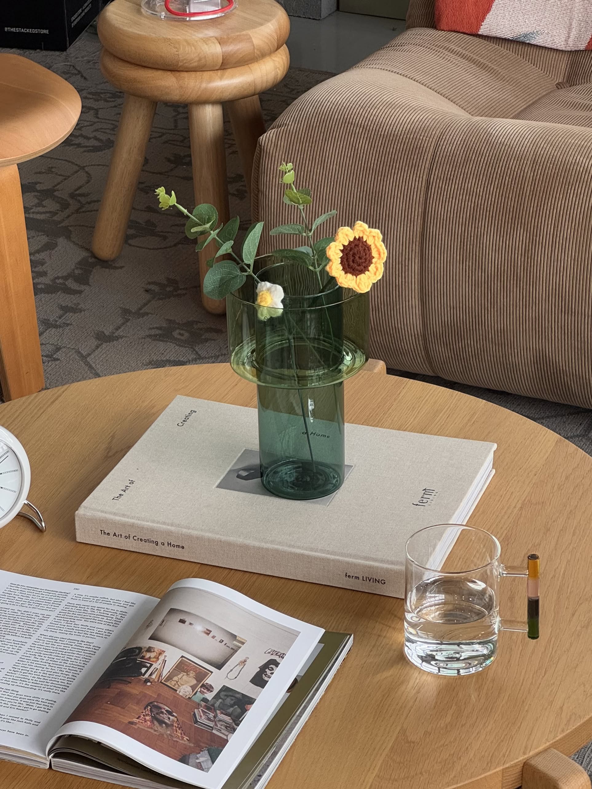The Ichendorf Milano Boreale Vase A, crafted from green borosilicate glass, features crocheted yellow flowers and sits on a large hardcover book atop a wooden table, adding artistic floral charm to a modern living room.