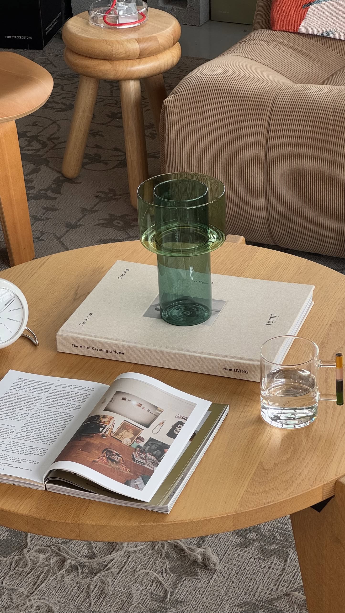 A wooden coffee table holds an open magazine, a closed book, a green Ichendorf Milano Boreale Vase A, and a glass of water. In the background, theres a beige couch and a patterned carpet.