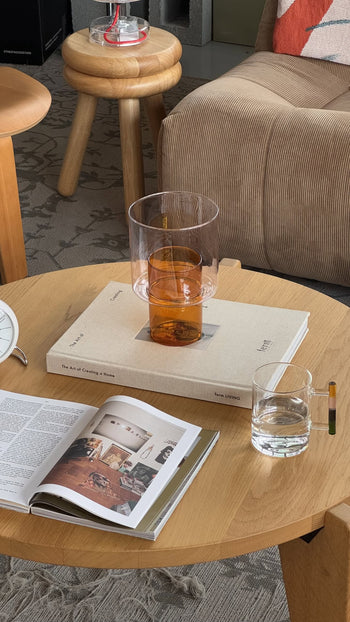 A wooden coffee table holds a beige book, the Ichendorf Milano Boreale Vase B in orange double-cylinder style, a clear glass of water, and an open interior design magazine. Nearby are a beige armchair and a patterned rug.