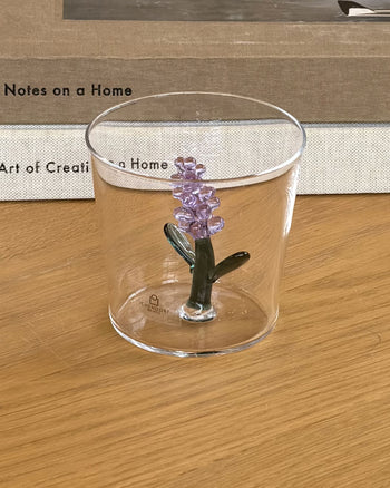 An Ichendorf Milano Botanica Tumbler - Lavender, featuring a purple flower design, sits on a light wooden table with a PROJECT JAPAN book and round white clock behind it, plus a sofa and red-patterned cushion in the background.