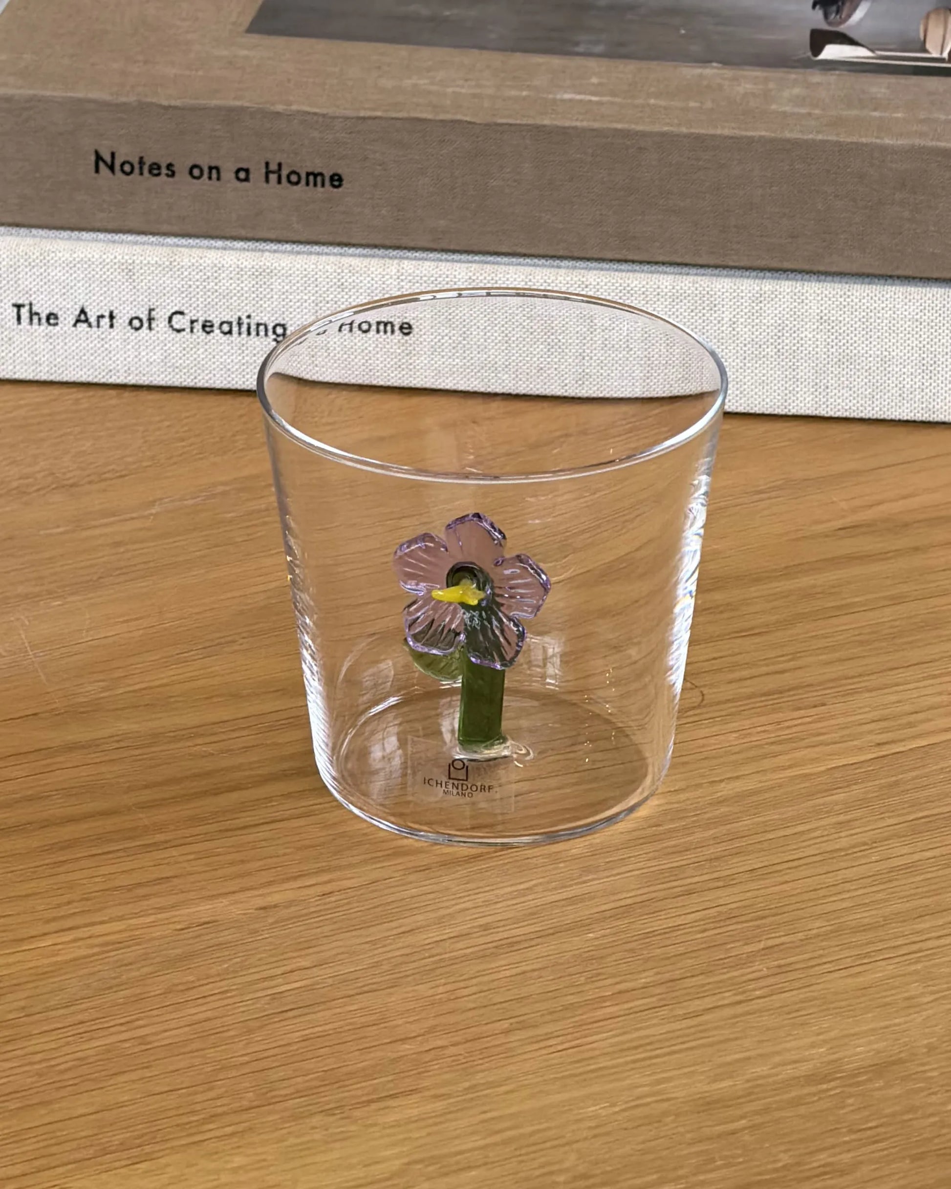 The Ichendorf Milano Botanica Tumbler - Lilac, featuring a delicate purple flower and green leaf motif, sits on a wooden surface, casting a clear shadow and adding nature-inspired charm to your space.