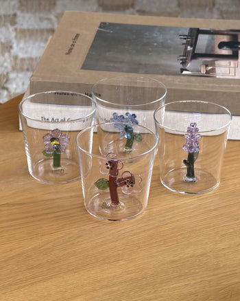 The Ichendorf Milano Botanica Tumbler - Lilac, a clear glass cup with a green stem-like handle and purple lilac flower by Ichendorf Milano, sits on a sunlit wooden surface, casting a charming nature-inspired shadow.