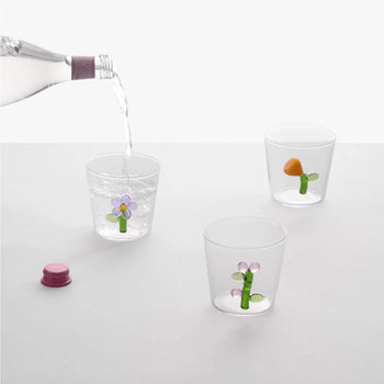 A hand pours water from a bottle into the Ichendorf Milano Botanica Tumbler - Lilac, its purple floral design standing out, while two more Ichendorf Milano cups from the Botanica collection display botanical and mushroom motifs on white.