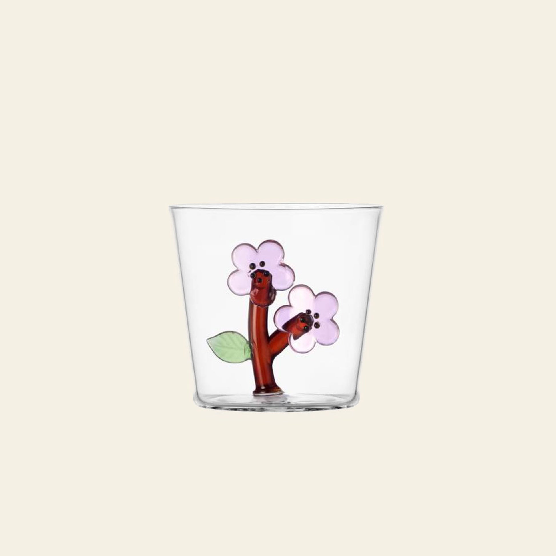 The Ichendorf Milano Botanica Tumbler - Peach Blossom is a clear glass tumbler featuring two cartoonish brown tree branches with smiling faces, pink flowers, and green leaves on a beige background. Brand: Ichendorf Milano.