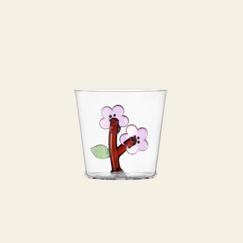The Ichendorf Milano Botanica Tumbler - Peach Blossom is a clear glass tumbler featuring two cartoonish brown tree branches with smiling faces, pink flowers, and green leaves on a beige background. Brand: Ichendorf Milano.